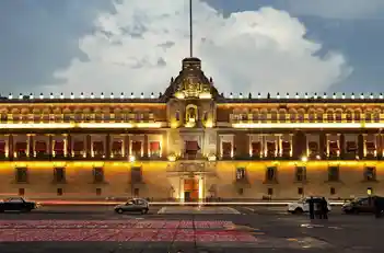Fachada del Palacio Nacional iluminada de noche frente a la Plaza de la Constitución.