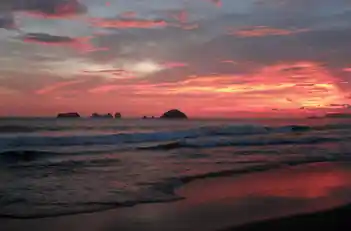 Atardecer con tonos rojizos y morados sobre las olas de Playa El Palmar e islotes al fondo.