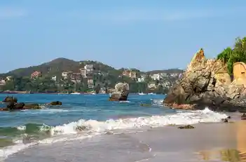 Oleaje suave en la orilla de Playa La Ropa con formación rocosa y colinas al fondo.