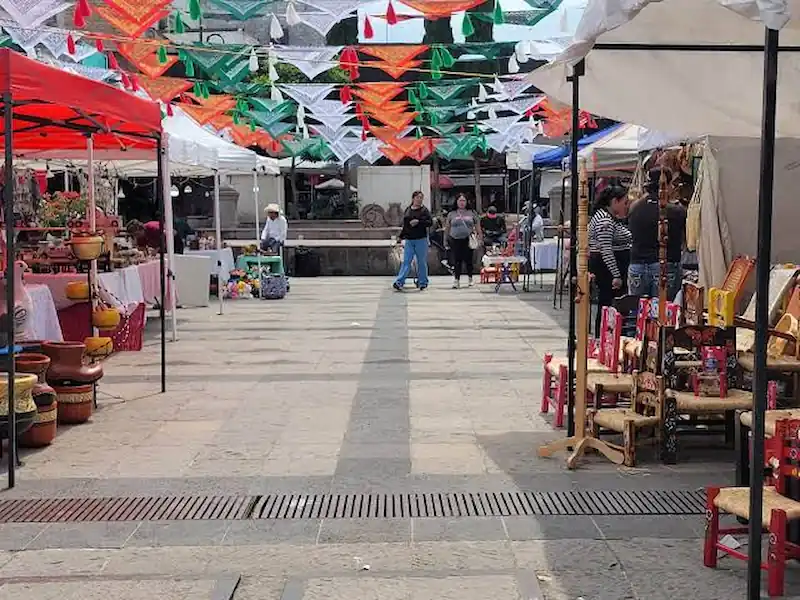 Pasillo al aire libre de un mercado artesanal con puestos bajo carpas, sillas de madera decoradas y papel picado multicolor.