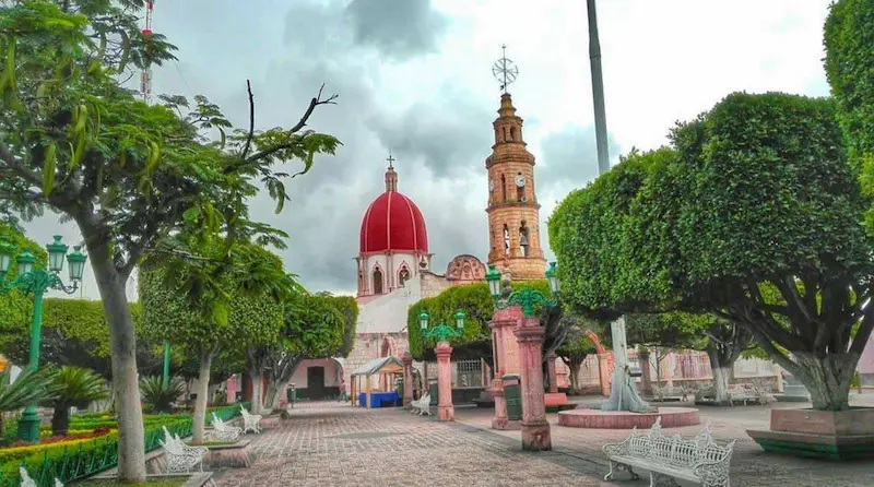 Plaza pública con bancas blancas, árboles podados y el Templo de Santiago Apóstol con su cúpula roja y torre al fondo.