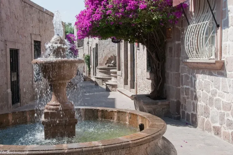 Fuente de cantera funcionando en un callejón colonial de Morelia adornado con bugambilias moradas.