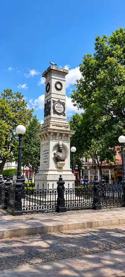 Monumento a Benito Juárez en la plaza principal de Uruapan rodeado de árboles y faroles tradicionales.