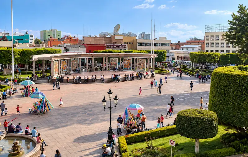 Vista aérea de la Plaza Cívica Benito Juárez en Zitácuaro con visitantes, arboledas y murales al fondo.