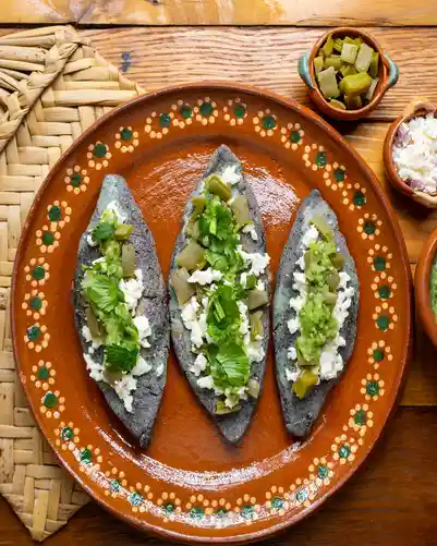Tlacoyos de maíz azul servidos en plato de barro con queso, nopales y cilantro.