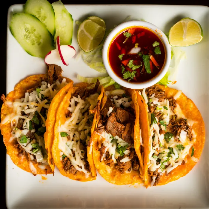 Cuatro tacos dorados de birria con queso fundido y cilantro servidos con consomé y rodajas de pepino y limón.