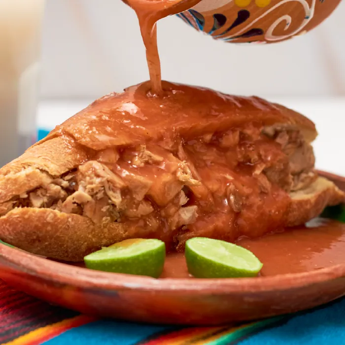 Torta ahogada en birote salado bañada con salsa de jitomate caliente sobre un plato de barro con limones.