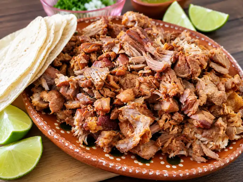 Platon de barro lleno de carnitas de cerdo picadas acompañado de tortillas de maíz y rodajas de limón fresco.