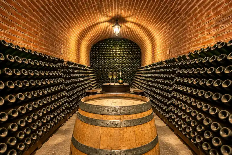 Cava subterránea de ladrillo con barrica de madera en primer plano y estantes repletos de botellas de vino.
