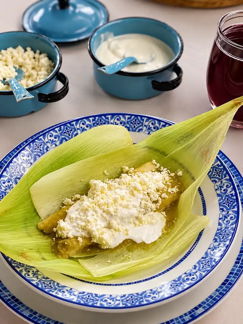 Uchepo de elote tierno servido en su hoja con salsa verde, crema y queso fresco rallado.